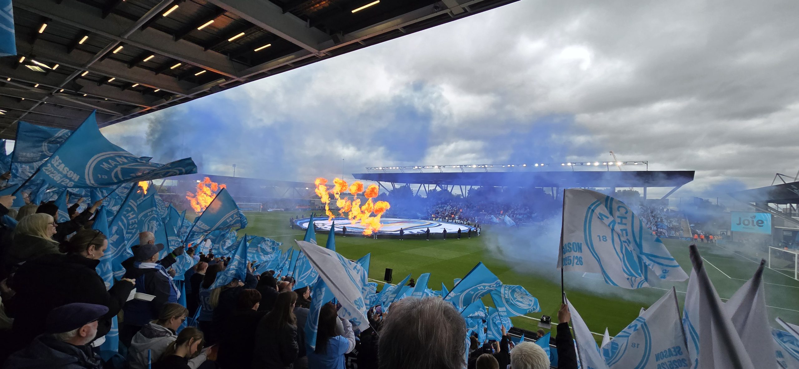 Photograph of the Joie Stadium before Mqanchester City vs Liverpool in May 2026, the sadium is full, blue smoke and flamethrowers are visible as flags are waved.